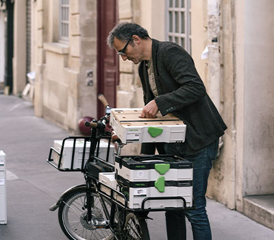 Nach getaner Arbeit packt der ungewöhnliche Tischler seine Werkstatt zusammen und macht sich mit dem Fahrrad wieder auf den Weg durch die Straßen von Paris.