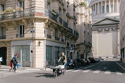 Mit seinem Lastenrad fährt der „ébéniste à vélo“ zügig durch die dichte Innenstadt von Paris zu einer Kundin, im Hintergrund die berühmte Kuppel des Panthéon.